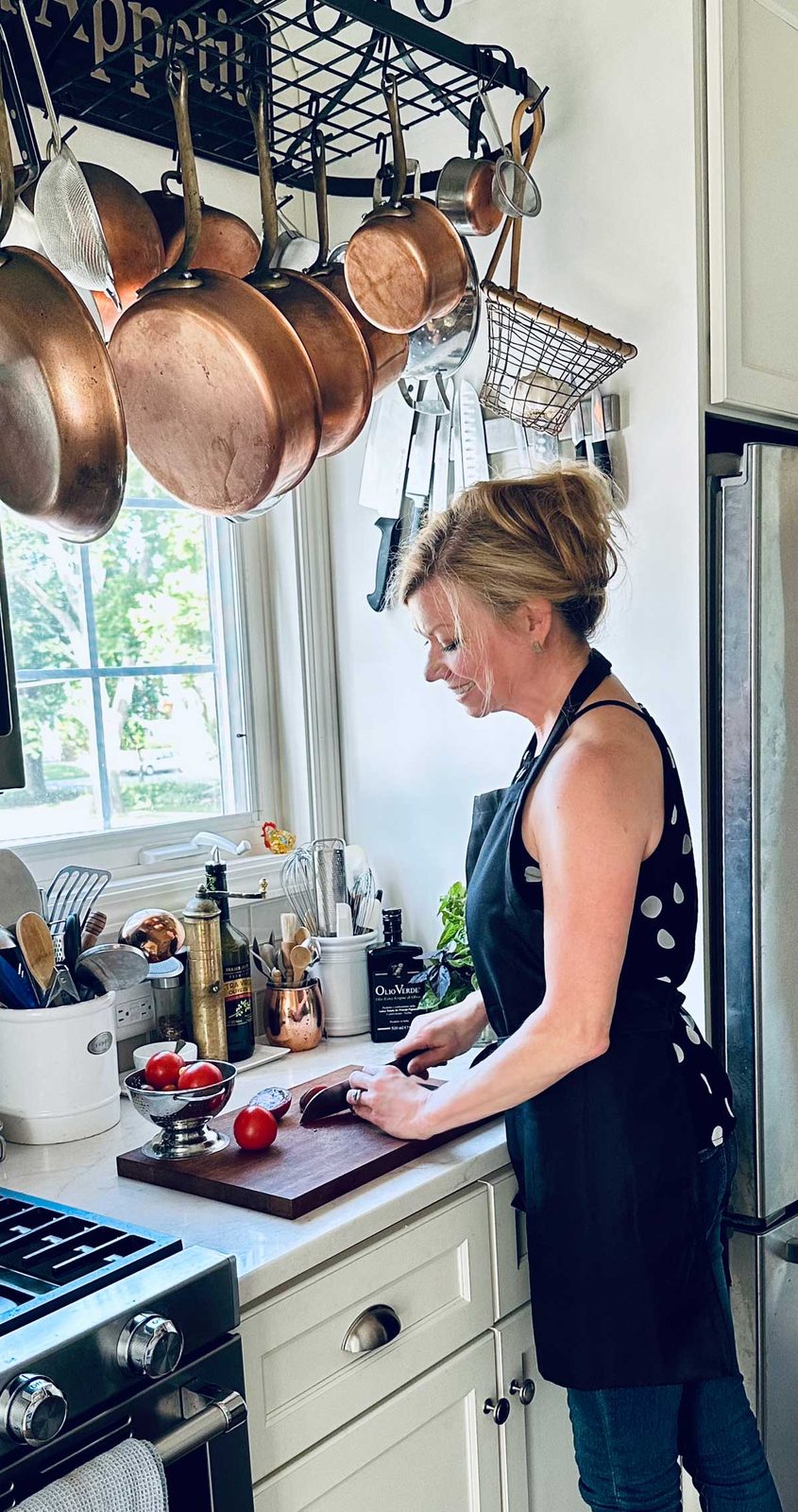 A woman wearing a black apron and polka-dot top chops tomatoes on a wooden cutting board in a bright kitchen with hanging copper pots and kitchen utensils. Sunlight streams through a window above the sink.