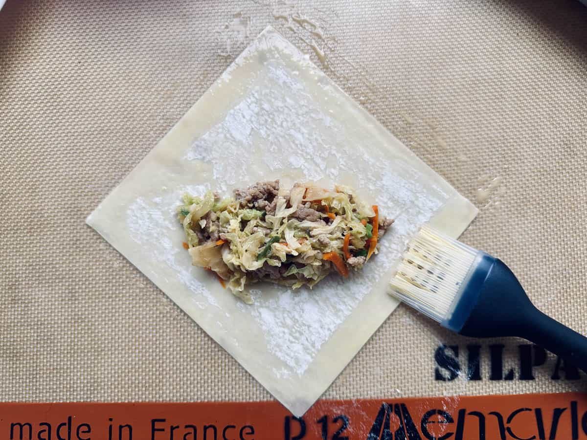 A square wrapper with a meat and vegetable filling is on a baking mat, next to a pastry brush. The filling includes shredded cabbage and carrots. The wrapper is dusted with flour, ready to be folded.