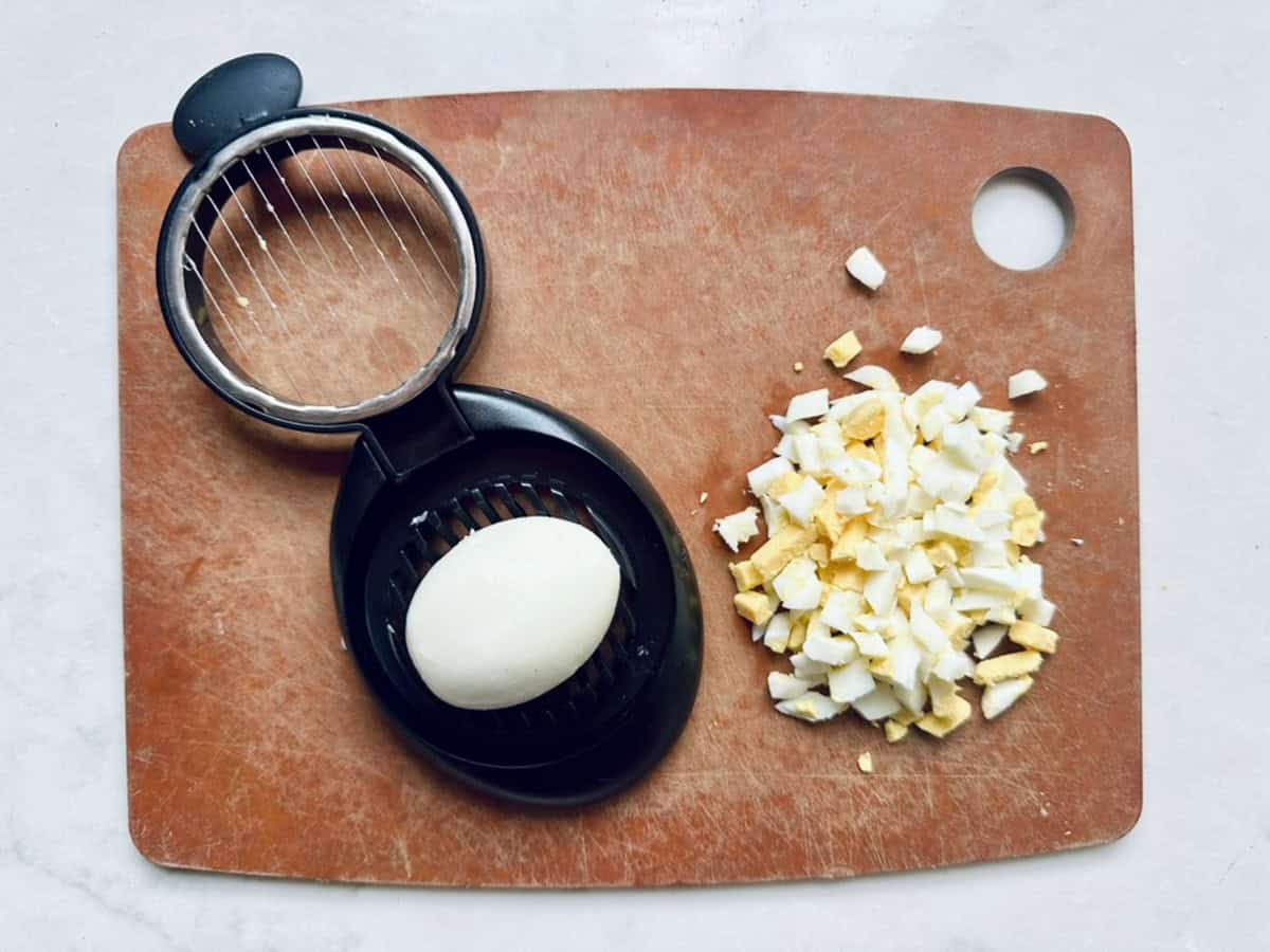 A hard-boiled egg sits in a black egg slicer on a brown cutting board. Next to it, chopped pieces of another hard-boiled egg are ready—perfect for making classic All-American Russet Potato Salad.
