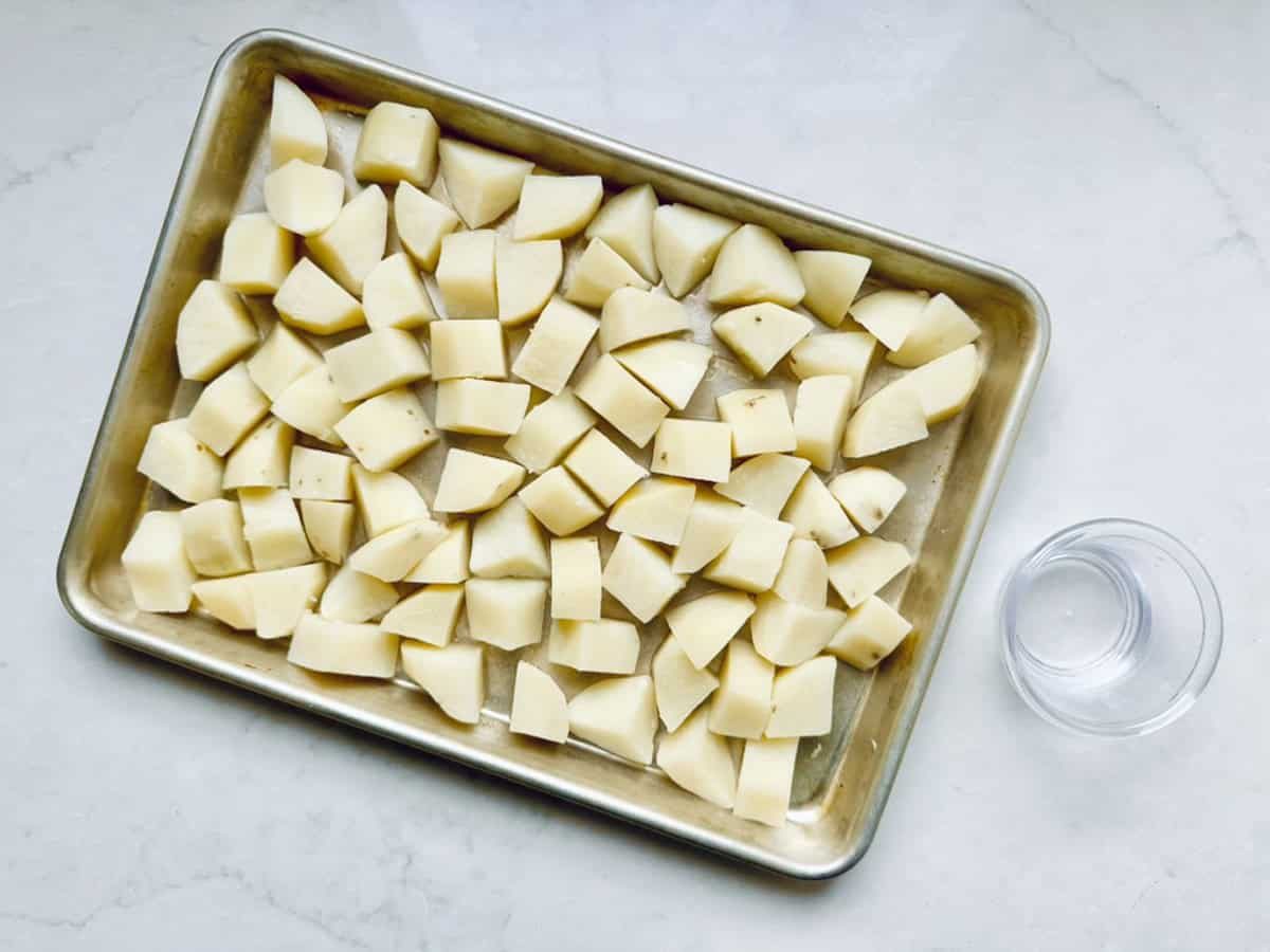 A baking tray filled with evenly spread, raw potato chunks—perfect for All-American Russet Potato Salad—sits on a light-colored countertop beside a small clear glass bowl of water.