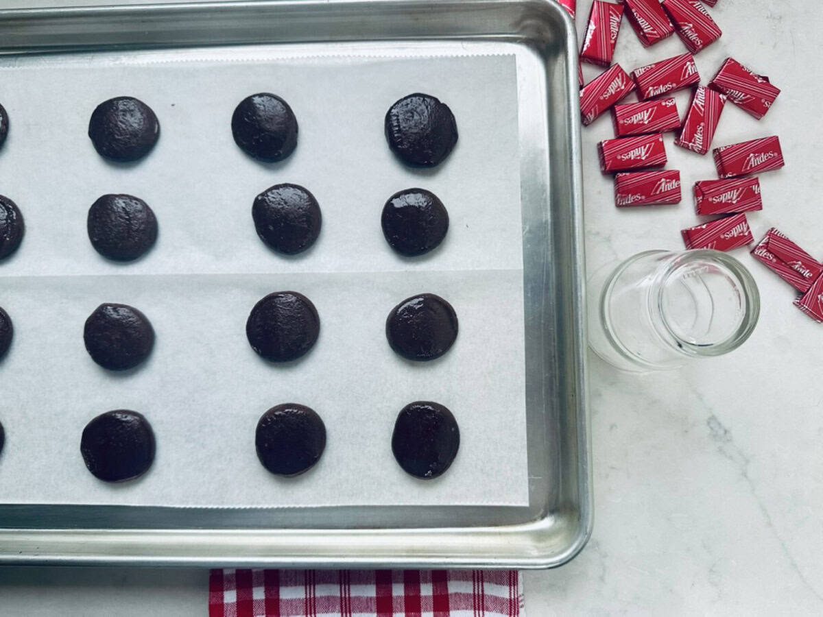 A baking sheet lined with parchment paper holds rows of unbaked chocolate cookie dough balls. To the right, red-wrapped Andes mints and a small empty glass jar sit on a white countertop with a red-striped cloth.