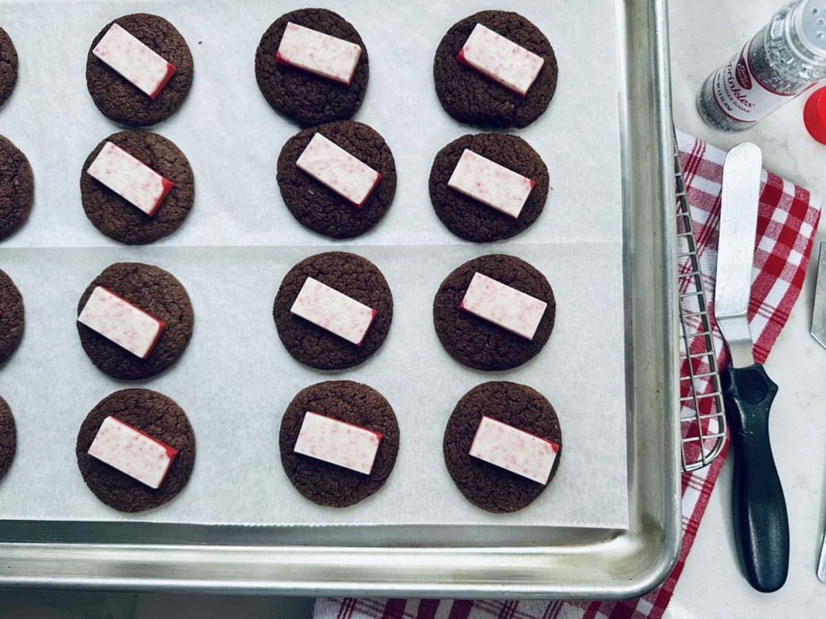 Rows of chocolate cookies on a baking sheet, each topped with a rectangular piece of peppermint bark. A red and white kitchen towel and a spatula are visible to the side.