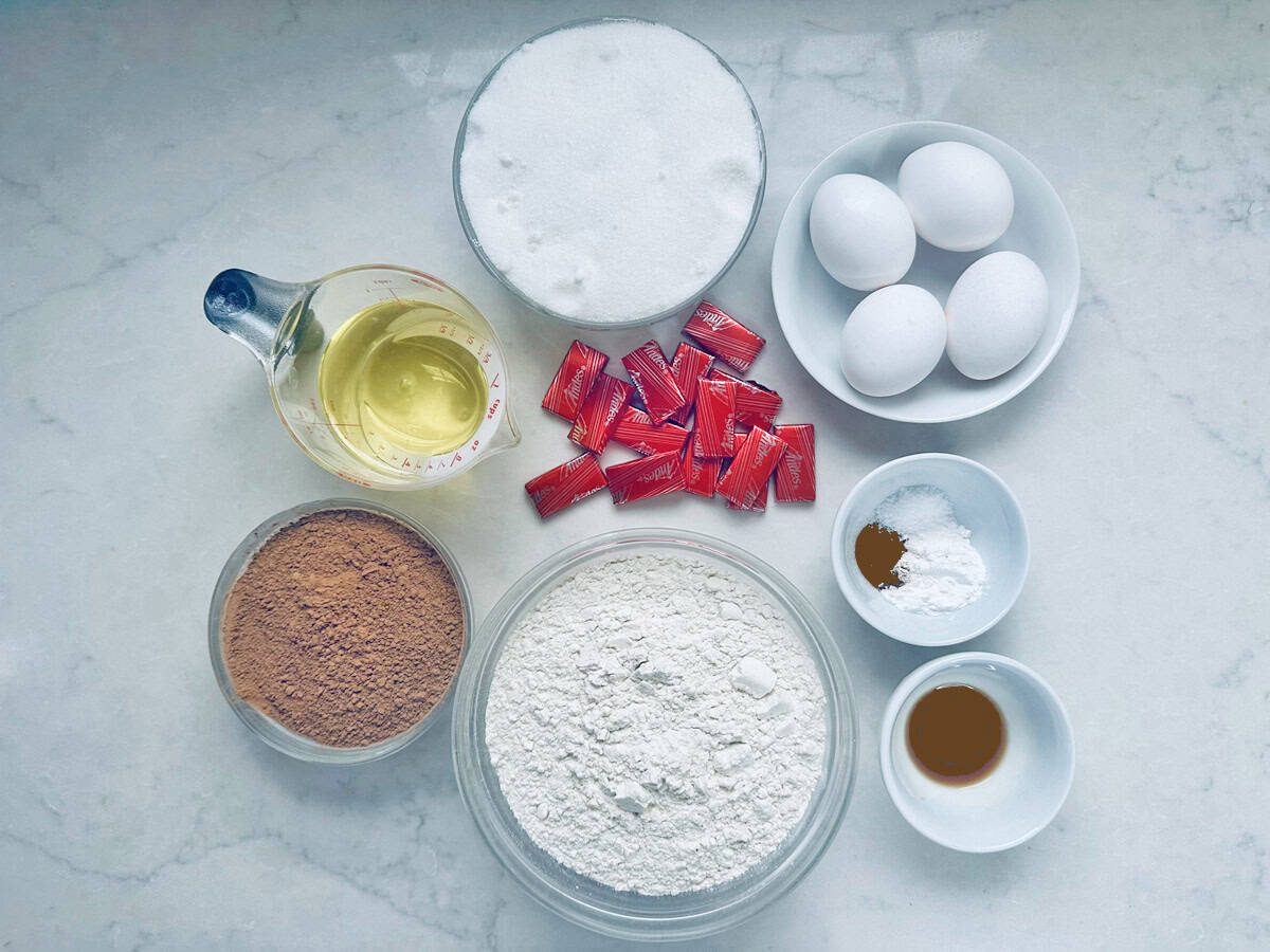 Baking ingredients on a counter: eggs, granulated sugar, cooking oil, flour, cocoa powder, red food coloring packets, baking powder, vanilla extract, and cinnamon, all in separate bowls and containers.