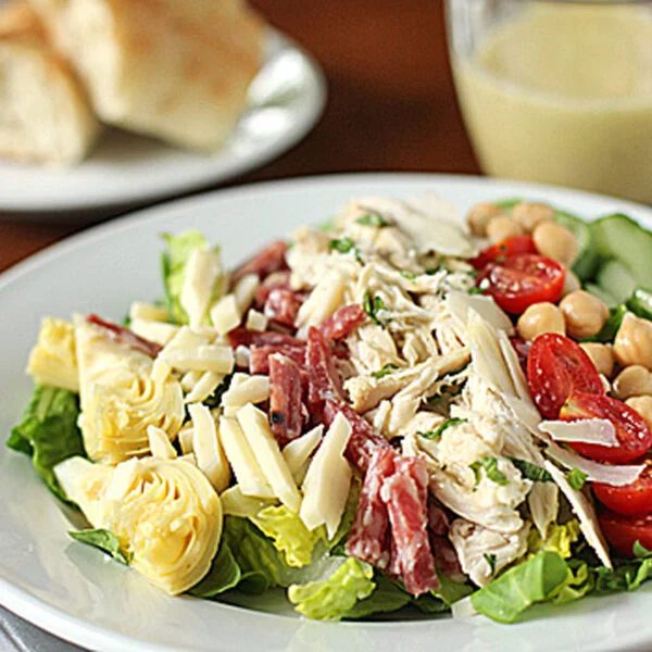 A plate of salad with sliced chicken, salami, artichoke hearts, cherry tomatoes, chickpeas, cheese, and lettuce. A fork and knife are beside the plate. Bread and a cup of dressing are in the background.