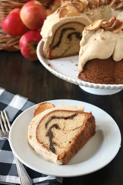 A slice of bundt cake with a cinnamon swirl and creamy frosting sits on a white plate, next to a fork. The rest of the cake, topped with dried apple slices, is displayed on a stand with apples in the background.