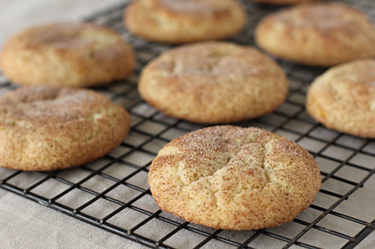 Freshly baked snickerdoodle cookies cooling on a black wire rack, topped with a light dusting of cinnamon sugar. The cookies appear soft, thick, and slightly cracked on top.
