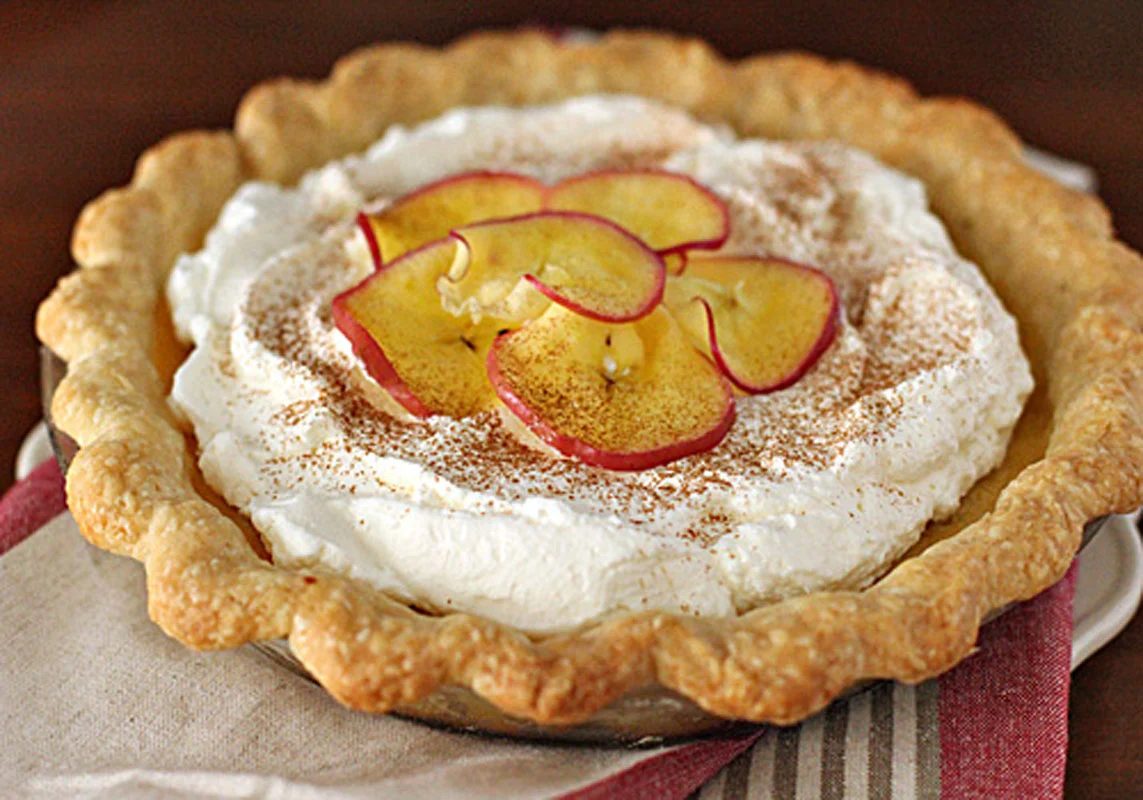 A pie with a golden, fluted crust, topped with fluffy whipped cream, cinnamon, and thin slices of apple arranged in a decorative pattern, sits on a folded napkin.