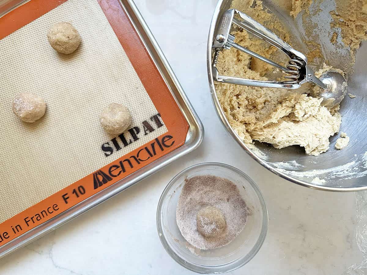 Apple Cider Snickerdoodles cookie dough balls being rolled in cinnamon sugar, placed on a baking sheet lined with a Silpat mat, with a bowl of dough and a cookie scoop nearby.