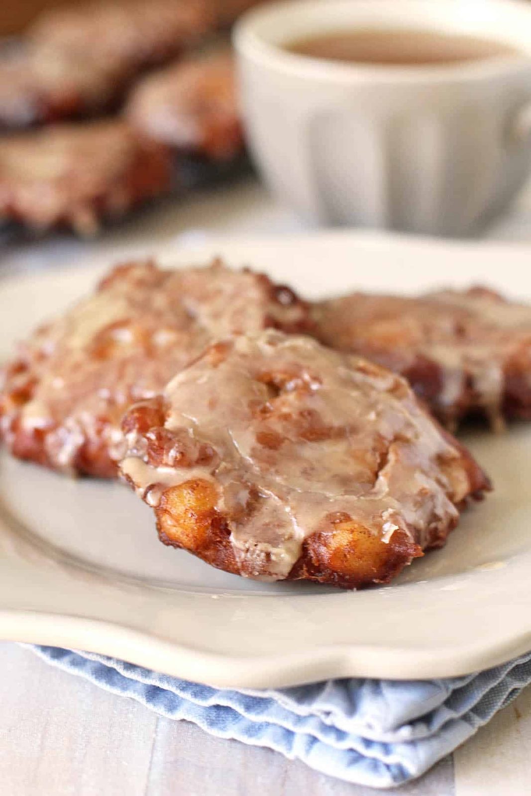 Two glazed apple fritters sit on a white plate with a cup of coffee in the background. The fritters are golden brown and topped with a shiny glaze. A folded blue napkin is under the plate.