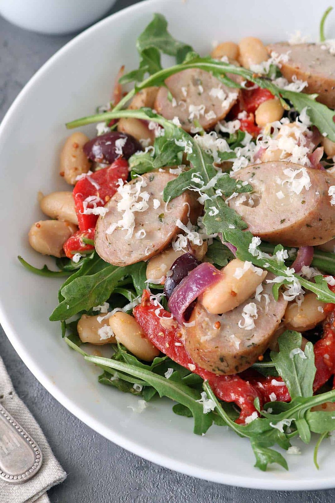 A bowl of salad with sliced sausage, arugula, white beans, roasted red peppers, olives, red onions, and grated cheese on top.
