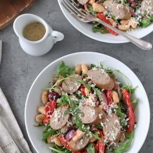 Two white bowls filled with salad featuring sliced sausage, white beans, arugula, red bell pepper, and grated cheese. A fork rests in one bowl. Nearby are sliced bread, a small jug of dressing, and napkins with cutlery.