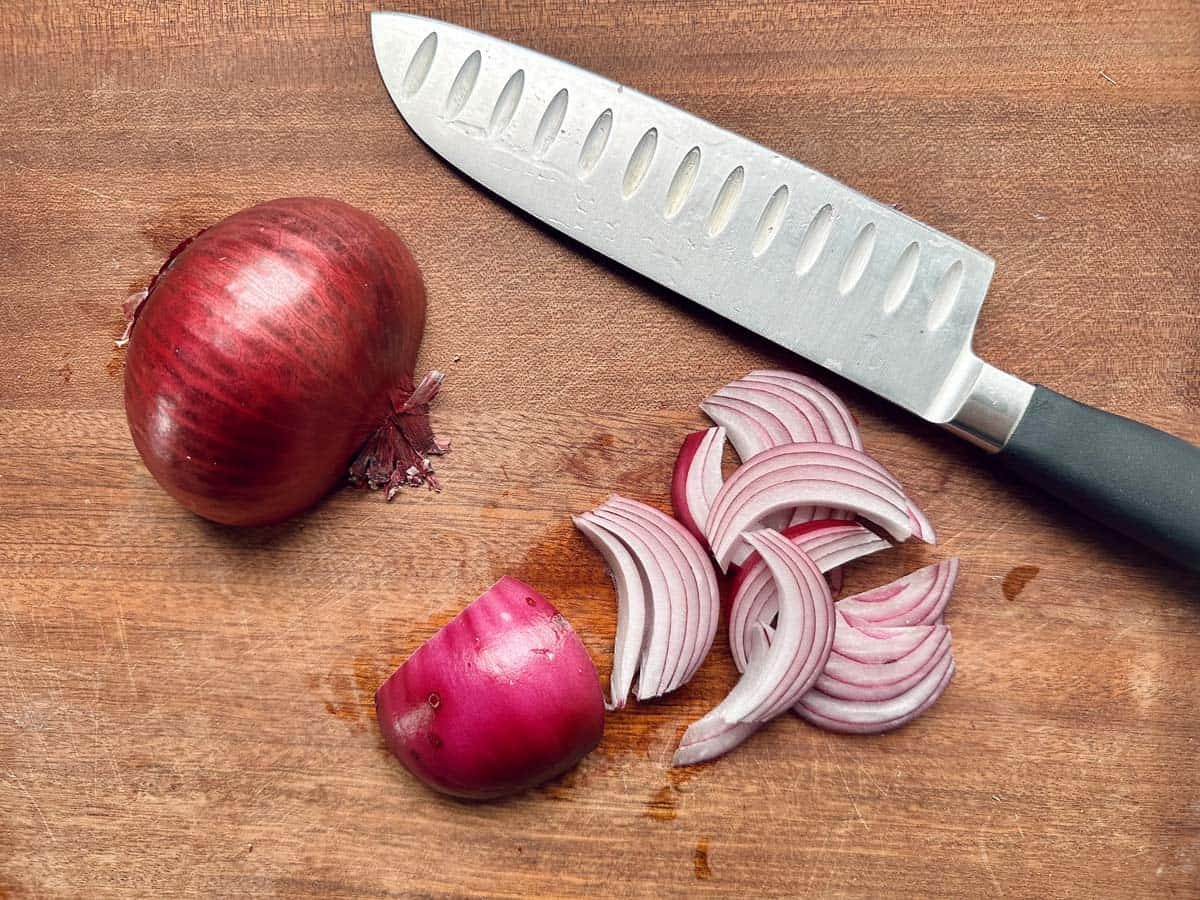 A halved red onion and several thinly sliced onion pieces sit on a wooden cutting board next to a large kitchen knife—ideal prep for arugula salad ingredients or quick salad meals.