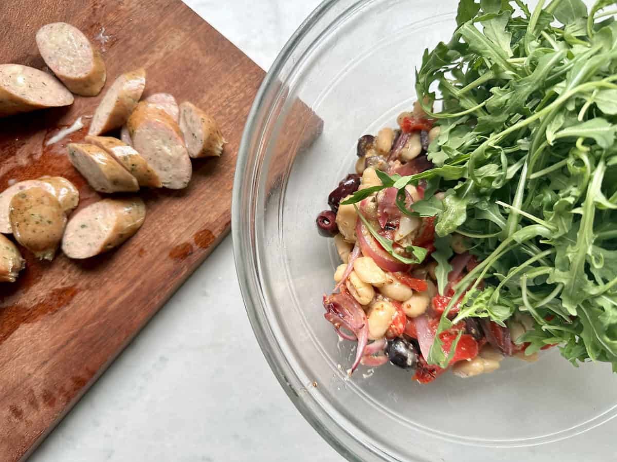 A glass bowl with arugula salad ingredients like beans, olives, and tomatoes, sits next to a wooden board with sliced sausage—perfect for sausage salad meals or an Arugula Salad with Sausage and White Beans.