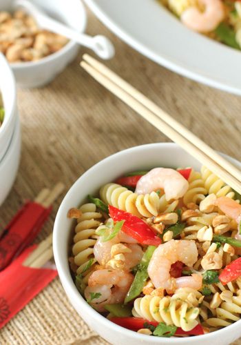 A bowl of pasta salad with shrimp, red bell peppers, snap peas, chopped peanuts, and herbs, served with chopsticks. A small bowl of chopped nuts and a napkin with chopsticks are visible nearby.