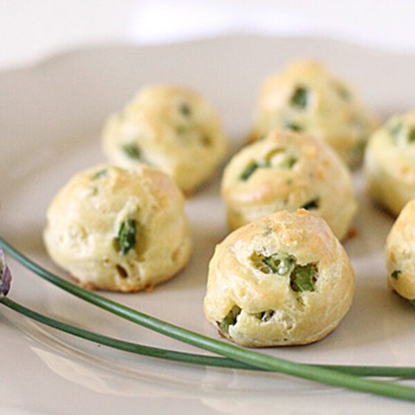A plate with several golden-brown, round pastry puffs with green herbs visible inside, displayed next to two purple chive flowers and green stems on a light-colored plate.