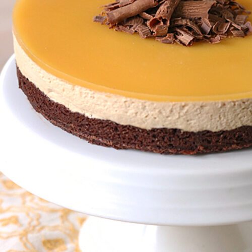 A layered mousse cake with a glossy caramel top, chocolate mousse middle, and brownie base, garnished with chocolate shavings, displayed on a white cake stand.