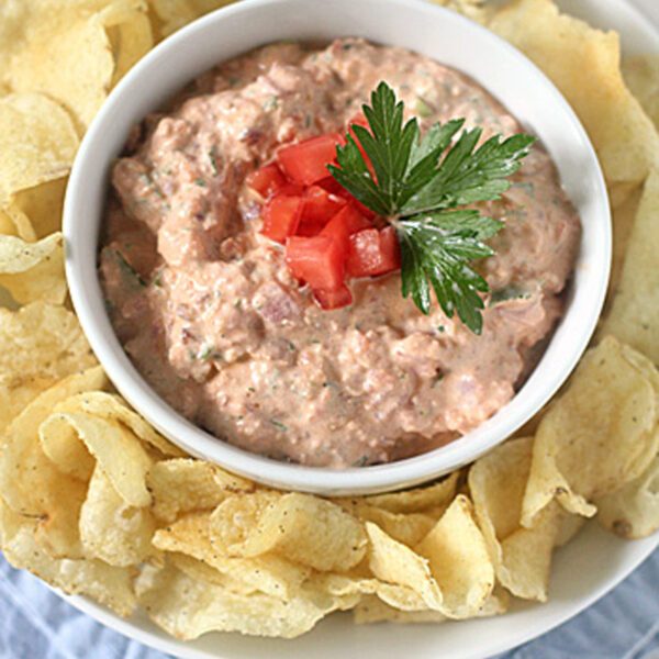 A bowl of creamy dip topped with chopped tomatoes and parsley, surrounded by potato chips on a white plate, set on a light blue napkin.