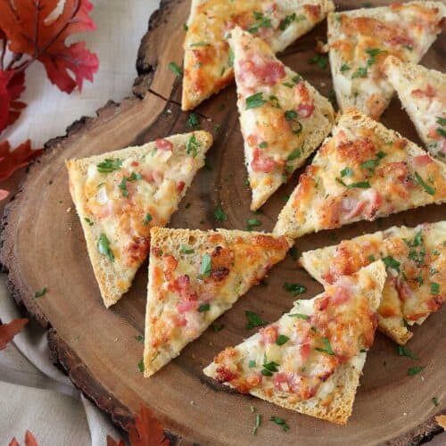 Slices of cheesy bread topped with bacon and herbs are arranged on a wooden serving board. Autumn leaves and a light cloth create a cozy, seasonal atmosphere around the board.