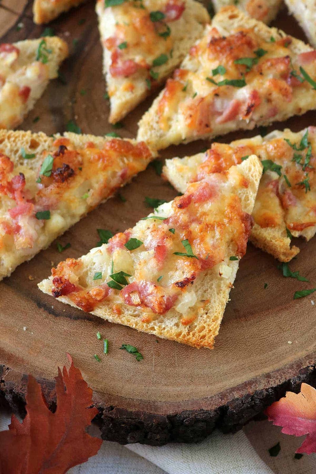 Several triangular slices of toasted bread topped with melted cheese, diced ham, onions, and fresh chopped herbs are arranged on a wooden serving board. Autumn leaves decorate the scene.