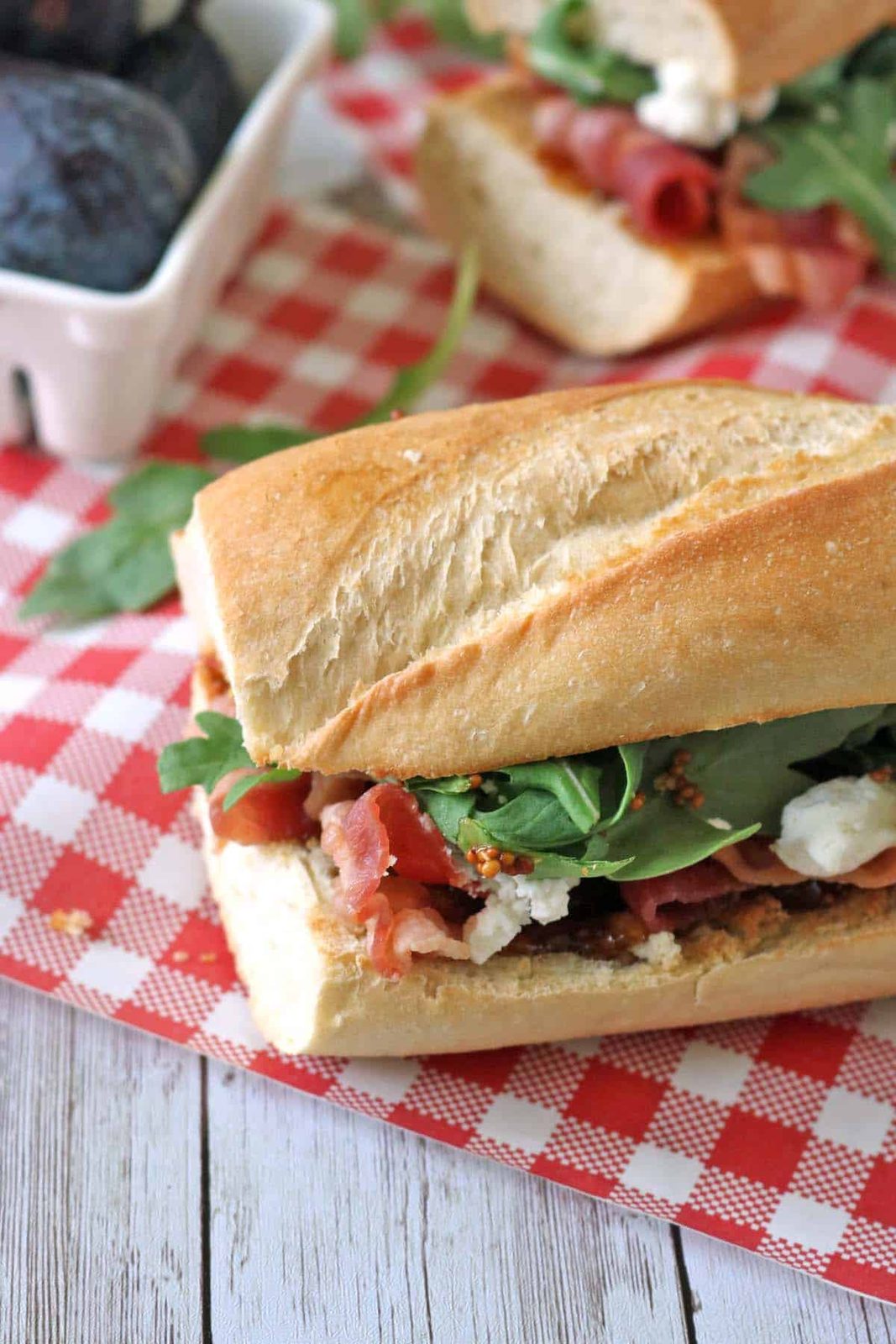 A sandwich with lettuce, prosciutto, and cheese on a baguette sits on a red and white checkered cloth, with a container of figs in the background.