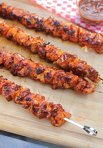 Four grilled chicken skewers with a glazed, slightly charred finish rest on a wooden cutting board, with a small bowl of sauce and red-checked paper visible in the background.