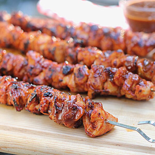 Four grilled meat skewers, coated in a shiny, reddish sauce, rest on a wooden cutting board. A small bowl of dipping sauce is visible in the background.