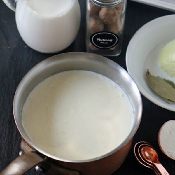 A saucepan filled with milk next to a small jug of milk, a jar of nutmeg, a white plate with onion and bay leaves, and copper measuring spoons on a dark surface.