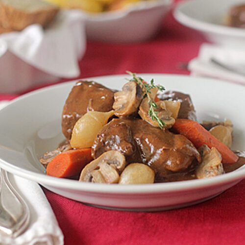 A white bowl filled with beef stew, carrots, mushrooms, and pearl onions, garnished with fresh thyme, sits on a red tablecloth next to silverware and a white napkin.