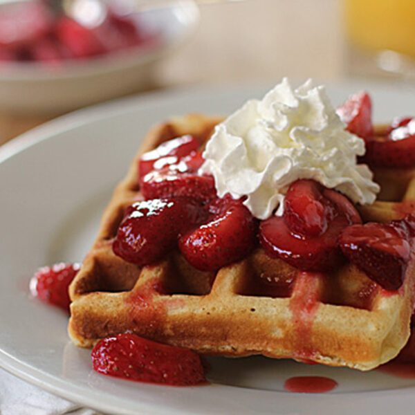 Two golden waffles topped with sliced strawberries, strawberry syrup, and a dollop of whipped cream are served on a white plate. A glass of orange juice and a bowl are blurred in the background.