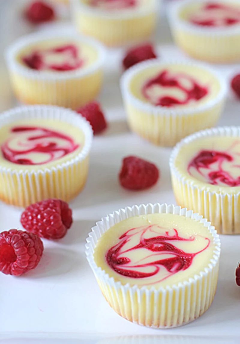 Mini cheesecakes in white cupcake liners, each topped with a red raspberry swirl, are arranged on a white surface with fresh raspberries scattered around them.