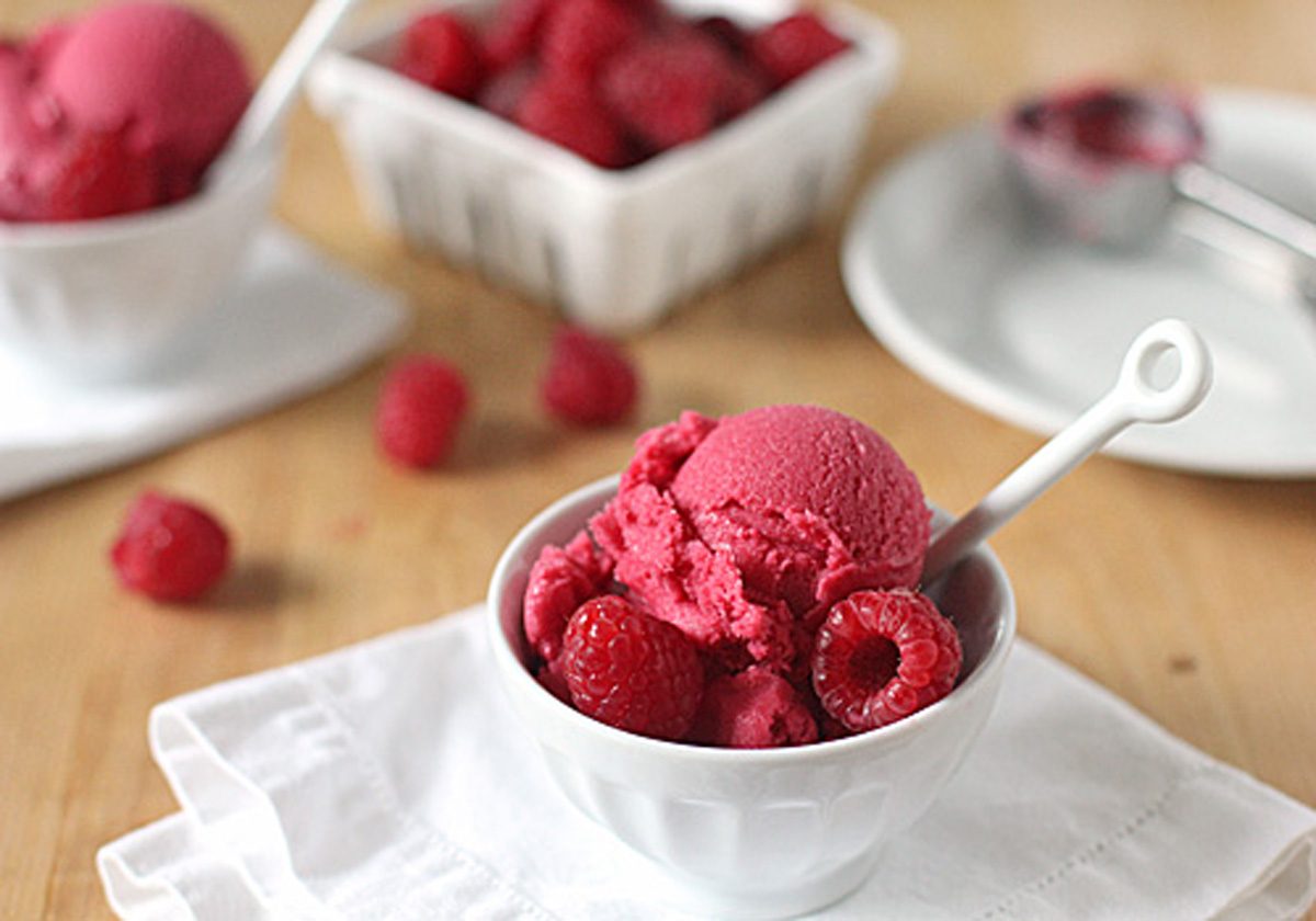 A white bowl filled with scoops of raspberry sorbet, garnished with fresh raspberries and a white spoon. In the background, there are more raspberries and a white container on a wooden table.