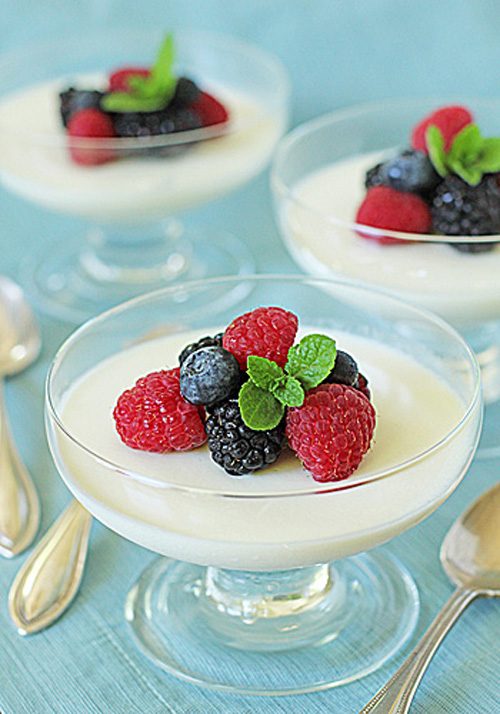 Glass dessert dishes filled with creamy panna cotta, each topped with fresh raspberries, blackberries, blueberries, and a small mint sprig, set on a light blue surface with silver spoons nearby.