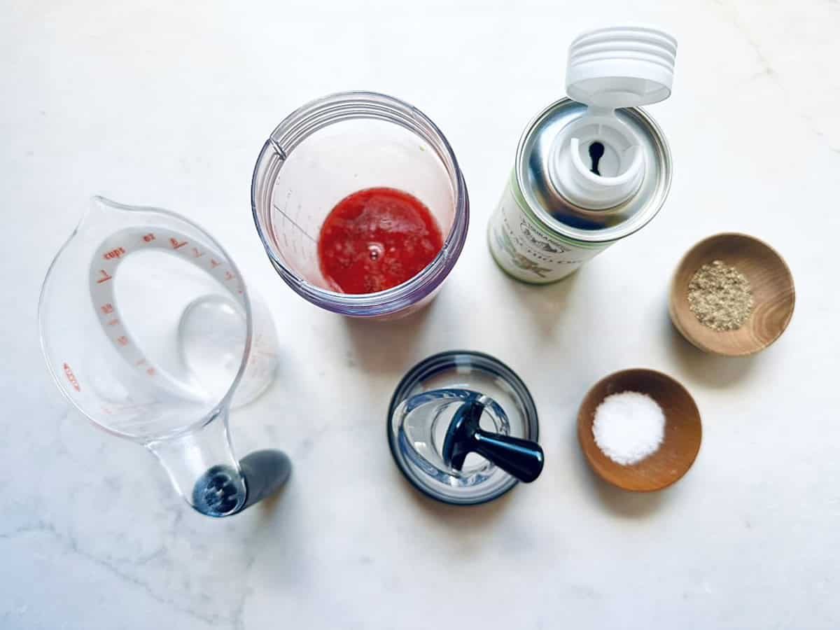 Top-down view of a measuring cup with water, a blender cup with red berry puree, an oil spray can, and three small bowls containing black pepper, salt, and white seasoning—perfect for making a Goat Cheese and Pistachio Salad.