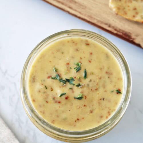 A glass jar filled with creamy mustard sauce garnished with fresh thyme, placed on a white marble surface near a wooden board and a spoon with sauce.