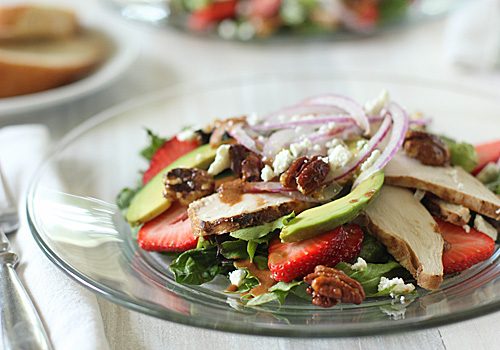 A glass plate with a salad of mixed greens, sliced strawberries, avocado, grilled chicken, red onion, crumbled cheese, and candied pecans, set on a white tablecloth with utensils and bread in the background.