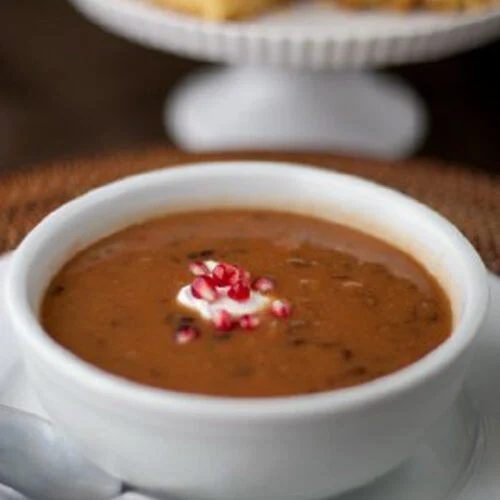 A bowl of bean soup topped with a dollop of cream and chopped red onions, served on a white plate with a spoon beside it; cornbread squares are displayed in the background on a cake stand.