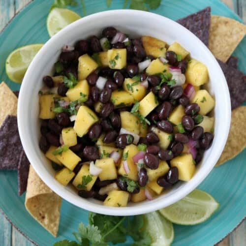 A bowl of black bean and mango salsa with chopped onions and cilantro, served on a blue plate with tortilla chips, lime wedges, and cilantro sprigs on a rustic wooden table.