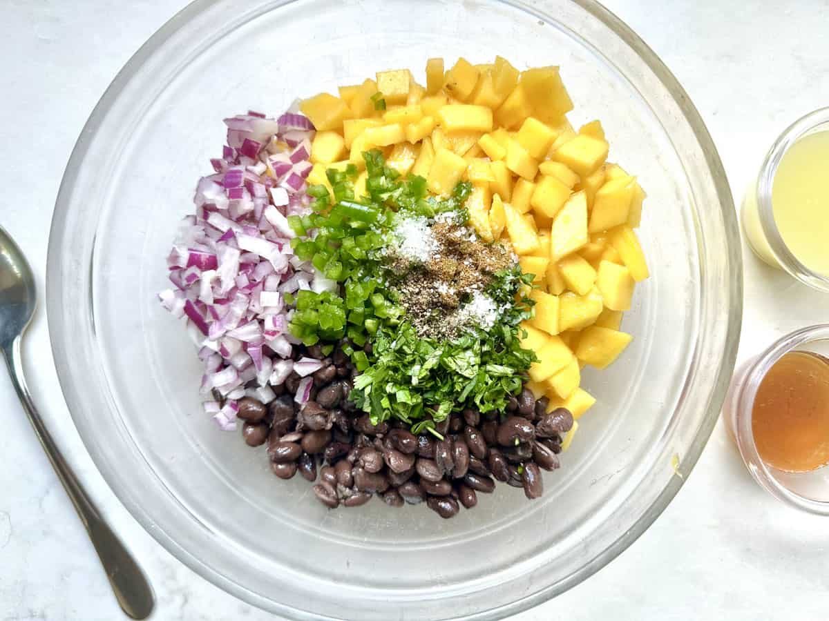 A glass bowl contains diced red onion, mango, jalapeño, black beans, chopped cilantro, and spices. Small bowls of dressing ingredients and a spoon are nearby on a white surface.