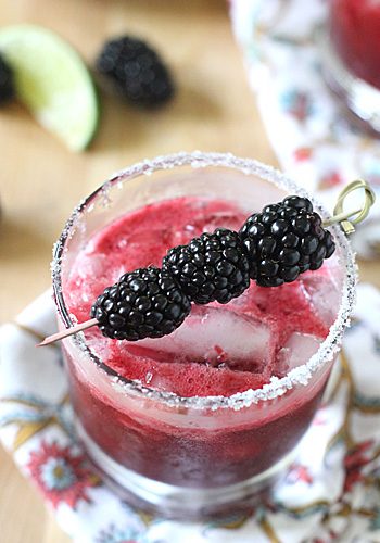 A glass filled with a purple blackberry cocktail on ice, rimmed with sugar, and garnished with three blackberries on a skewer. A lime wedge and blackberries are blurred in the background.
