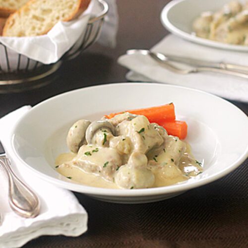 A white bowl filled with creamy chicken and mushroom stew, garnished with parsley and served with whole cooked carrots. A basket of bread and a plate with cutlery are beside the main dish on a dark table.