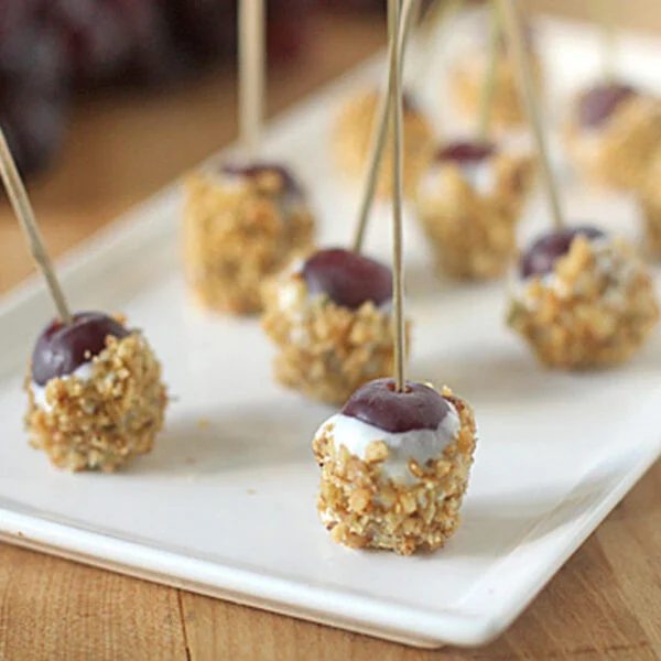 A rectangular white plate with several grape bites, each coated in a crumbly mixture and pierced with a toothpick, arranged neatly on a wooden surface. Grapes and more snacks are blurred in the background.