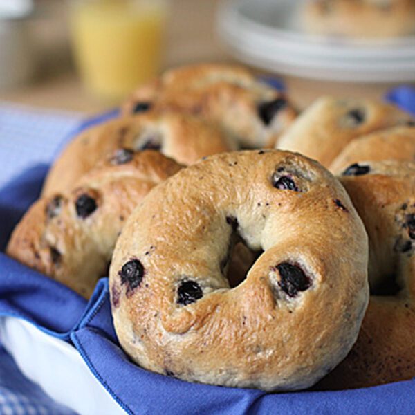 A basket lined with a blue cloth holds several freshly baked blueberry bagels, with more plates and a glass of orange juice blurred in the background.