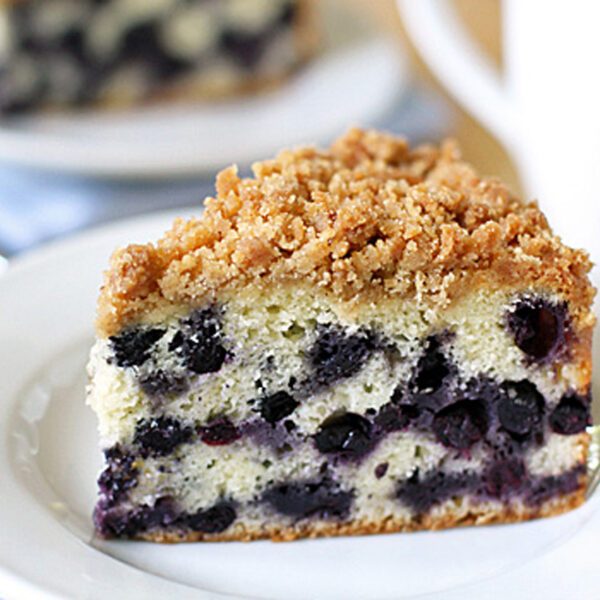A slice of blueberry crumb cake with streusel topping sits on a white plate. The cake has a light, fluffy texture with visible blueberries throughout. A blurred cup and another cake slice are in the background.