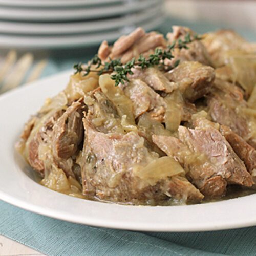 A white plate filled with tender, cooked pork pieces and sliced onions in a light sauce, garnished with a sprig of fresh thyme, set on a blue placemat with stacked plates and cutlery in the background.