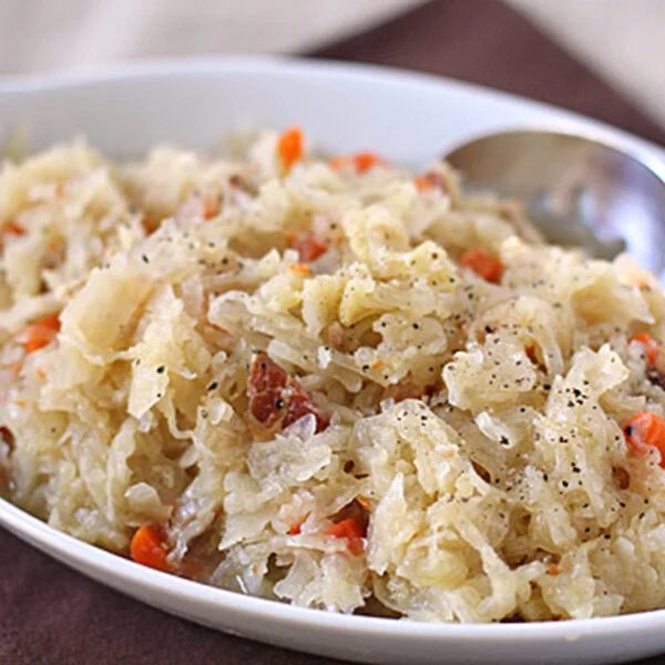 A close-up of a white dish filled with sauerkraut mixed with shredded carrots and small pieces of meat, garnished with black pepper, and a spoon resting on the side.