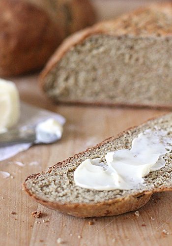A slice of brown bread with a layer of butter on top sits on a wooden surface, with a butter knife and a partially sliced loaf of bread in the background.