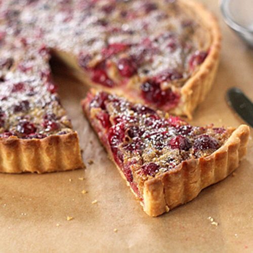 A cranberry tart with a golden crust sits on parchment paper. One slice is cut and slightly separated, showing the berry filling. A knife and a mesh strainer with powdered sugar are nearby.