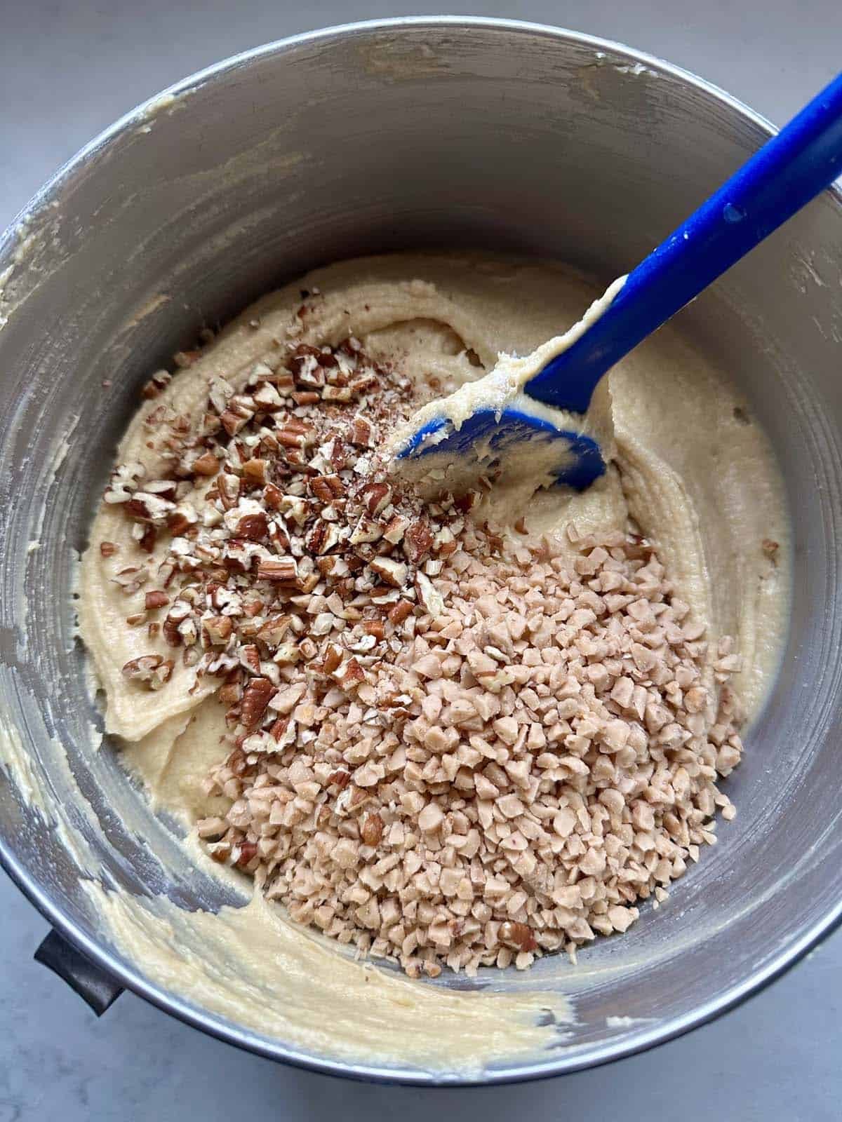 A mixing bowl with cookie dough being stirred, topped with chopped pecans and toffee bits, and a blue spatula resting in the batter.