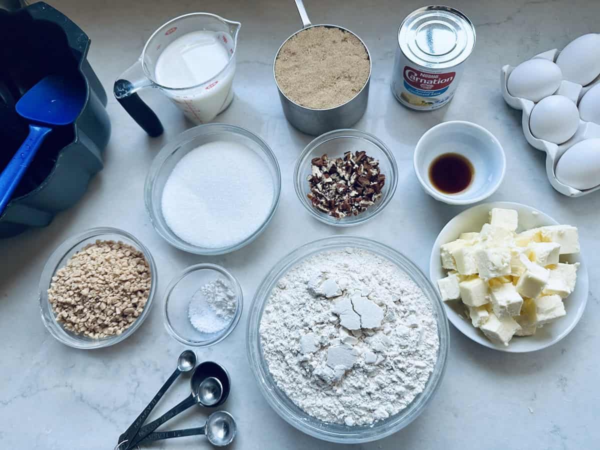 Baking ingredients arranged on a white countertop, including flour, butter, eggs, vanilla, pecans, brown sugar, granulated sugar, evaporated milk, baking powder, salt, measuring spoons, and blue spatulas.