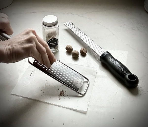 A hand grates whole nutmeg on a metal grater over a sheet of paper—perfect for adding fresh spice to a brown sugar pound cake. Nearby sit a nutmeg jar, three whole nutmegs, and another grater on a white countertop.